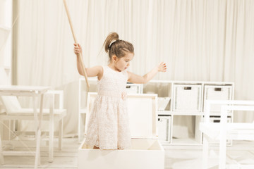 Happy little girl playing indoor pretending to fish in white wooden box boat with a stick as fishing line. Children imagination or creativity concept.