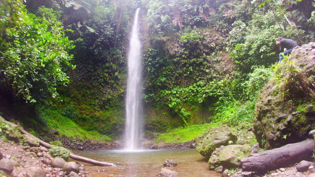 A serene tropical waterfall cascades into a shallow pool amid dense green foliage and moss-covered rocks, a person sitting on a boulder observing - the Hola Vida falls in the Ecuadorian Amazon.