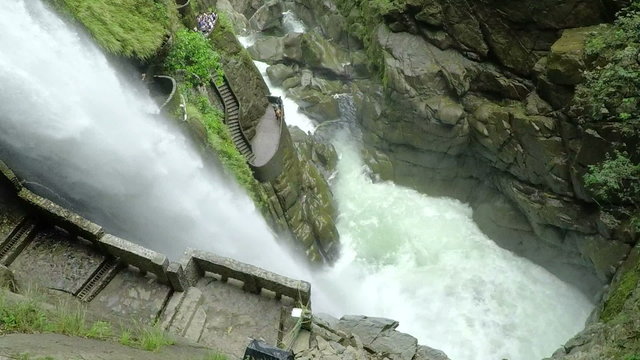 A high-angle view of the Pailon del Diablo waterfall plunging into a turbulent pool near Banos, Ecuador, a stone staircase winding along the cliffside as visitors watch from above.
