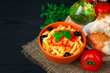 fusilli pasta and olives with parsley and ketchup in a bowl near to the tomatoes and parsley with chunks of fresh bread on a wooden black background