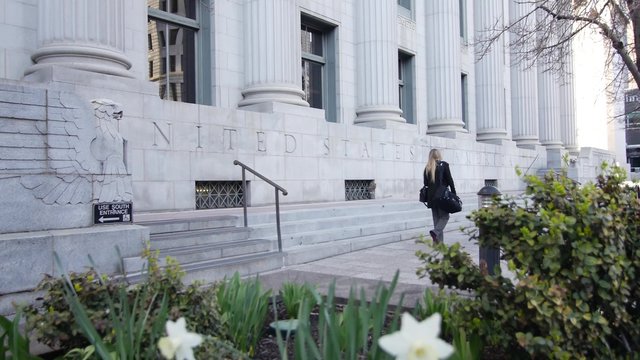 A woman strolls past blooming flowers at the foot of a stone courthouse. 
