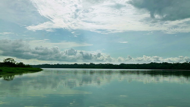 A serene wide shot of a tranquil river reflecting a dynamic cloudy sky, bordered by lush green forested banks, from a moving boat on Limoncocha lagoon in the Ecuadorian Amazon.