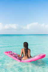 Obraz premium Beach vacation woman relaxing on ocean water bed. Beautiful woman from the back sitting on a pink pool float air mattress looking at view of the perfect turquoise pristine sea in tropical destination.