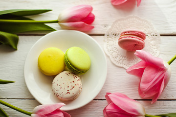Obraz na płótnie Canvas A beautiful flowers pink tulips with colorful macaroons laid on a white platter on white wooden background