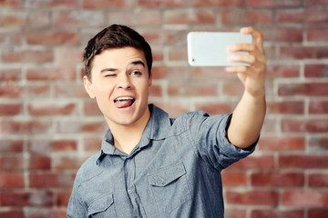 Young handsome boy making photo by his self with mobile phone on brick wall background