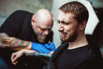 Fototapeta premium master tattooist makes a tattoo on the skin. The client narrows his eyes and turns away, the concept of pain and fear