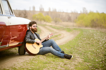 Obraz premium Bearded man playing guitar outdoors near retro car