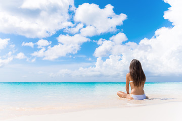 Beach vacation dream woman enjoying summer holiday on dreamy perfect ocean tropical destination. Person sitting from the back alone on deserted white sand beach getaway.