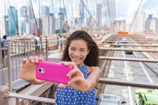 Happy New York City selfie tourist young woman taking a self-portrait photo with smartphone app on Brooklyn Bridge, NYC, Manhattan, USA. Asian girl doing mobile phone photography for social media.