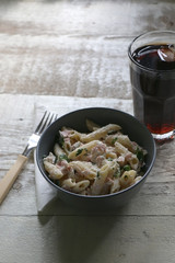 Pasta penne is cooked al dente, then mixed with sliced ham, feta cheese and basil. Dressing is made with olive oil, bit of vinegar, salt and pepper. Served with glass of soda on white rustic table.