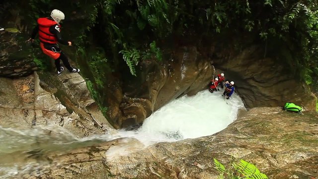 A group of adventurers in safety gear rappel into a turbulent waterfall pool in a lush jungle canyon, one descending as others wait on the rocks, near Banos, Ecuador.