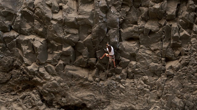 A rock climber ascends a steep, jagged cliff face while belayed from below, progressing up the rugged dark rock formation with a river at the base, in Ecuador.