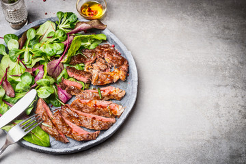 Sliced grilled beef steak with green leaves salad on rustic plate with cutlery. Medium rare...