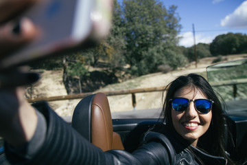 Woman smiling taking a selfie with her phone in a convertible car