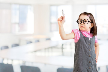 Cute little girl wearing business dress and draws something on virtual screen. Office background.