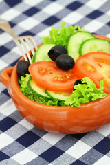 Bowl of salad consisting of lettuce, tomatoes, cucumber and black olives on checkered cloth
