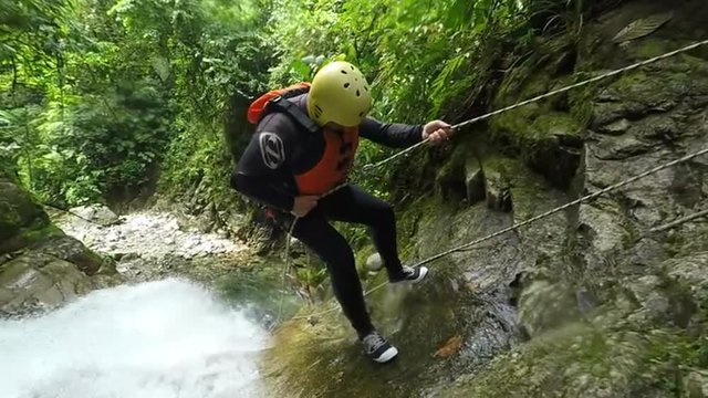 A man in a yellow helmet and orange life vest descends a mossy rock face beside a rushing jungle stream using fixed ropes, lush green forest around the narrow ravine near Banos, Ecuador.
