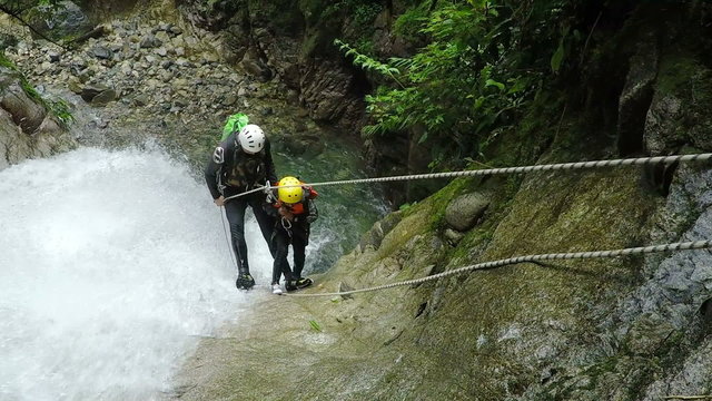 Two canyoneers in helmets and harnesses navigate a rocky streambed beside a rushing waterfall using fixed ropes in a lush green canyon, canyoning in the Ecuadorian Andes near Banos.