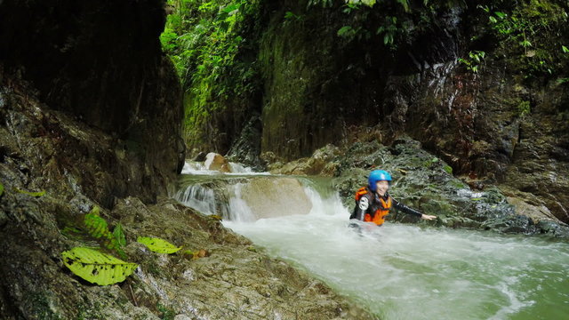 A person in a blue helmet and orange life vest stands on a rock in a rushing jungle stream amid moss-covered rocks and lush green foliage, canyoning in Llanganates National Park, Ecuador.