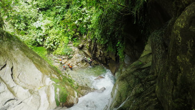 A first-person view of a canyoneer stepping down a mossy rock face above a rushing river, other adventurers below in a lush green canyon in Llanganates National Park, Ecuador.