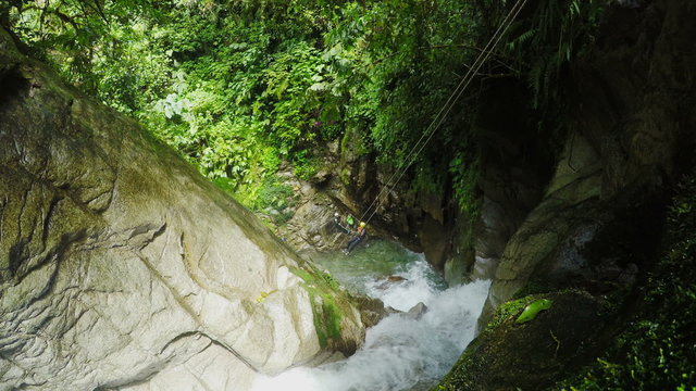 A high-angle view of adventurers rappelling down a narrow moss-covered river gorge amid dense tropical vegetation, turbulent water below, in Llanganates National Park, Ecuador.