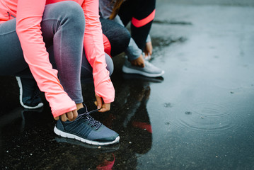 Urban athletes lacing sport footwear for running over asphalt under the rain. Two women getting...