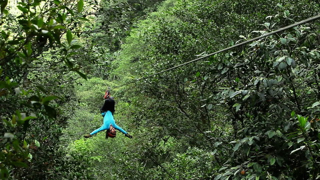 A woman in athletic wear ziplines through dense green jungle near Banos, Ecuador, transitioning from seated to an inverted spread-eagle pose, shot from a low angle among the foliage.