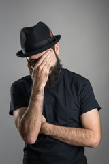 Facepalm of bearded hipster wearing black hat and t-shirt.  Headshot portrait over gray studio background with vignette. 