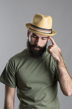 Worried thinking bearded man wearing straw hat looking down and scratching hand with finger. Portrait over gray studio background. 