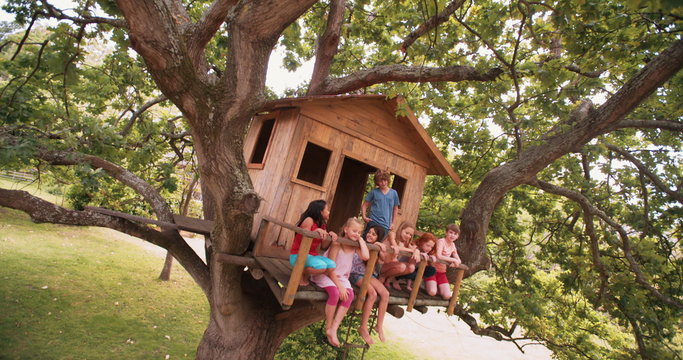 Children sitting on edge of treehouse in green leafed tree