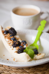 Banana dessert with granola, organic blueberries and fresh mint on a white plate with light green spoon. Coffee with healthy sweet dessert for snack.