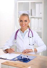 Beautiful young smiling female doctor sitting at the desk and writing.