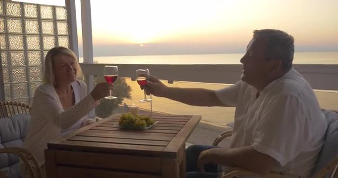 Senior man and woman enjoying time face to face. They having red wine relaxing in chairs on the outdoor balcony  at sunset. Romantic evening