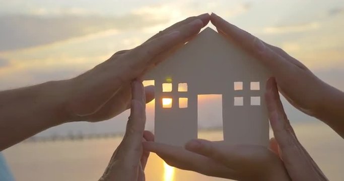Close-up shot of woman holding home model and then family making house around it with hands. Sunset and sea as background. Zooming in to get the sun in the window