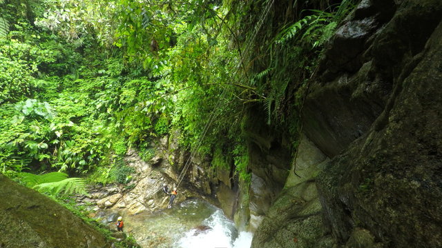 A person in an orange harness rappels down a mossy rock face in a lush tropical canyon near Banos, Ecuador, a river below and another adventurer assisting the descent.