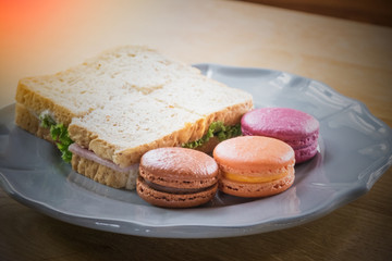 Abstract of colorful macaroon and sandwich ham cheese ready to eat / Abstract of orange, blueberry and chocolate macaroon with ham cheese whole wheat bread sandwich in ceramic plate on wooden table