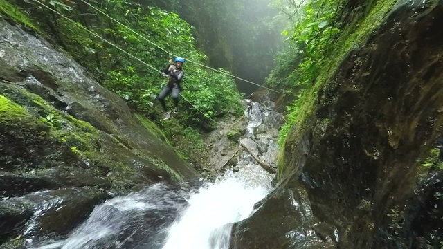 A canyoneer in safety gear crosses a rope bridge over a rushing waterfall in a lush moss-covered jungle canyon in Llanganates National Park, Ecuador, a wide view of the steep rock walls.