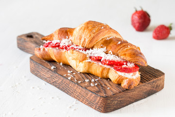 Croissant with cream cheese, strawberries and coconut flakes on wooden cutting board on white background. Healthy light breakfast table setting
