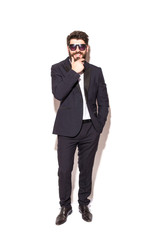 Full height Cheerful young handsome man in sunglasses keeping hand on chin and looking at camera with smile while standing against white background