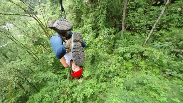 A woman in a red helmet ziplines through dense tropical jungle near Banos, Ecuador, smiling as she glides over green foliage and a rocky riverbed in a high-angle tracking shot.