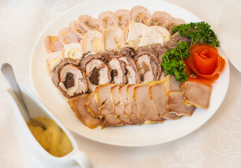 Close up of white plate full of meat appetizers decorated with red rose made of tomato and fresh leaves of parsley. Assortment of sliced meat, chicken and pork rolls. Saucepan with sauce.