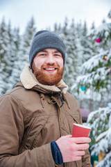 Man drinking coffee near decorated christmas tree in winter forest