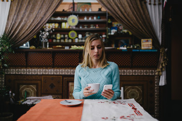 Portrait of a cute blonde female read something on her smart phone while sitting in ethnic coffee shop beautiful young hipster girl using cell phone while enjoying her free time in expensive cafe