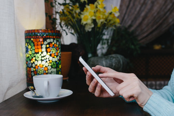 Woman typing text message on smart phone in a cafe on the wooden table, in cafe, near the window . Cropped image of young woman sitting at a table with a coffee using mobile phone.