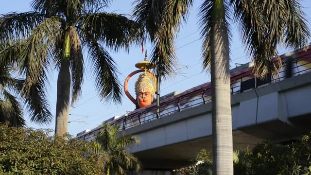 Locked-on shot of metro train moving in front of giant statue of lord Hanuman, Karol Bagh, New Delhi, India 