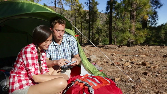 Camping couple in tent using smartphone looking at pictures photos. Campers smiling happy outdoors in forest. Happy multiracial couple having fun in outdoor activity. Asian woman, Caucasian man