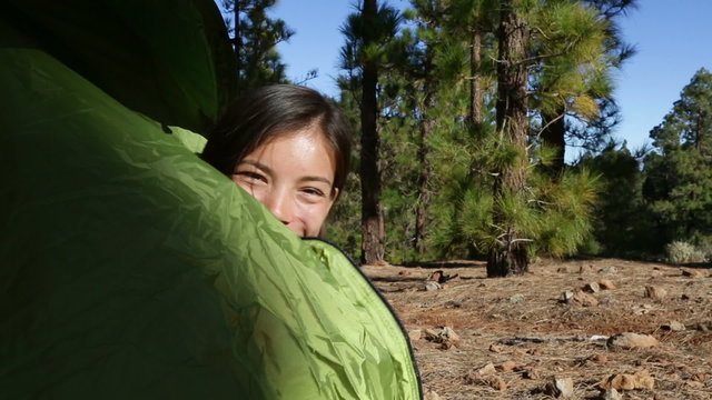 Woman camping waving hello from tent smiling happy outdoors in forest. Happy girl saying hello and goodbye closing tent. Smiling mixed race Asian Caucasian girl saying hi looking at camera.