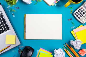 Office table desk with set of colorful supplies, white blank note pad, cup, pen, pc, crumpled paper, flower on blue background. Top view and copy space for text