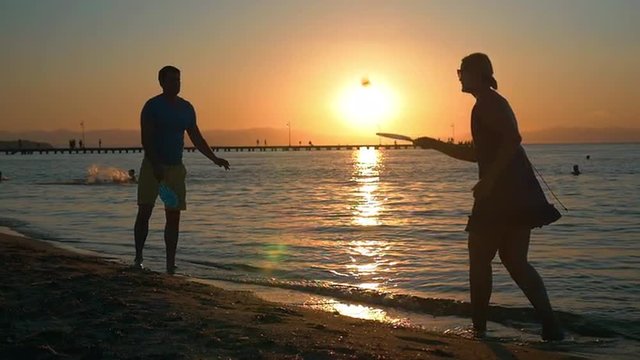 Slow motion of young man and woman playing tennis by the sea at sunset. Golden sun reflecting in dark water. Active and carefree summer vacation