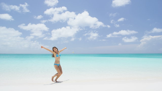 Happy bikini woman having fun jumping of joy and happiness on beach splashing water in perfect turquoise water. Asian girl cheering winning for Caribbean vacation destination during summer travel.